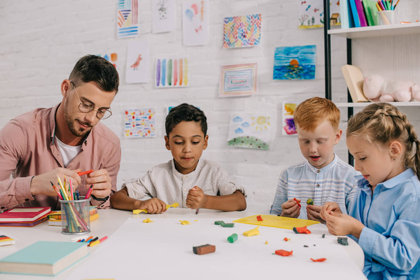 multiracial preschoolers and teacher with plasticine sculpturing figures at table in classroom - Photo, Image