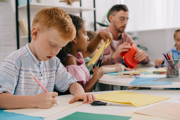 szelektív összpontosít, tanár és többnemzetiségű óvodás gyerekek színes papírokkal, hogy papír Applied osztályteremben  - Fotó, kép