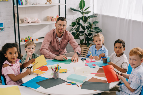 педагог мужского пола и многорасовые дошкольники сидят за столом с разноцветными докладами в классе
 - Фото, изображение