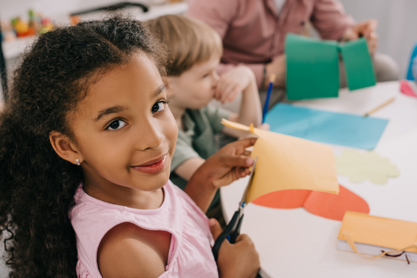 enfoque selectivo de preescolares multiétnicos cortando papeles coloridos con tijeras en el aula
  - Foto, Imagen