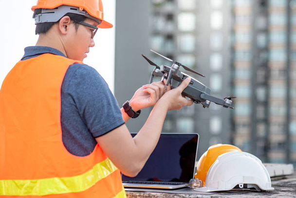 Nuori aasialainen insinööri mies työskentelee drone kannettavan tietokoneen ja älypuhelimen kanssa rakennustyömaalla. Miehittämättömän ilma-aluksen (UAV) käyttö maa- ja rakennustyömaan kartoitukseen maa- ja vesirakennushankkeessa
. - Valokuva, kuva