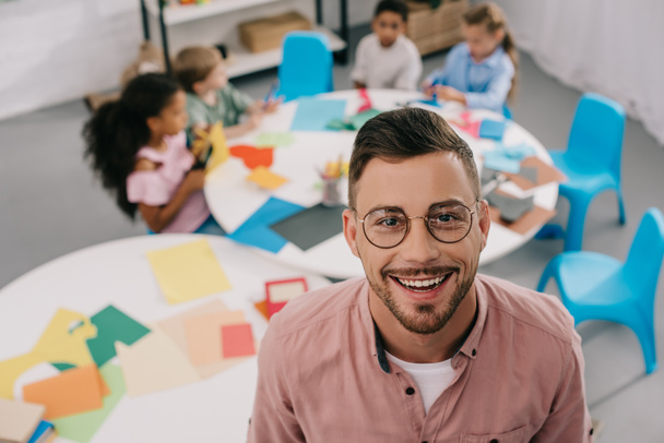 enfoque selectivo del profesor sonriente en anteojos y niños interracial en la mesa en el aula
 - Foto, imagen