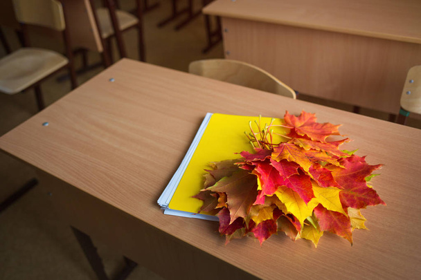 School classroom in blur background without young student. notebook and bouquet of autumn maple leaves on the table. concept: back to school, teacher's day - Photo, image