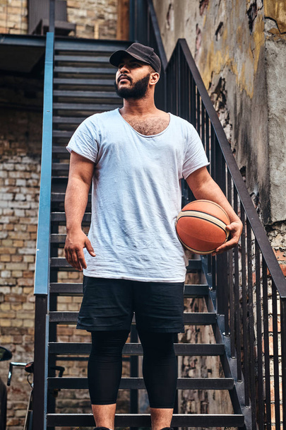 Portrait of an African-American bearded guy in cap dressed in a white t-shirt and sports shorts holds a basketball standing on stairs in a ghetto, looking away. - Foto, afbeelding