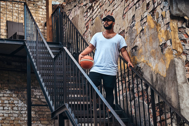 Портрет афроамериканского бородатого парня в кепке, одетого в белую футболку и спортивные шорты, держит баскетбольный мяч, стоящий на лестнице в гетто, отворачиваясь
. - Фото, изображение