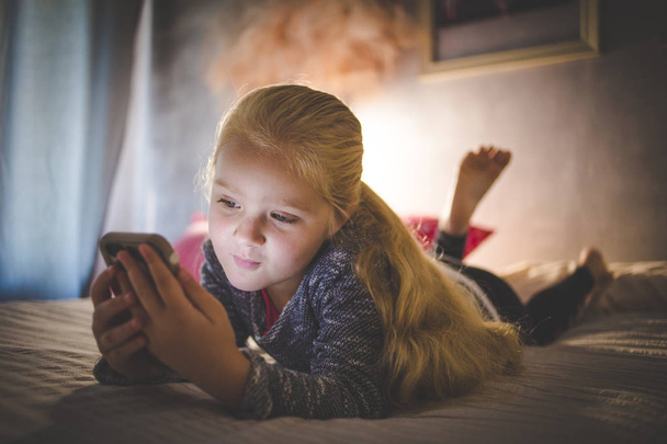 Piękne blond toddler dziewczynka r. na jej łóżku oglądając filmy na smartfonie - Zdjęcie, obraz