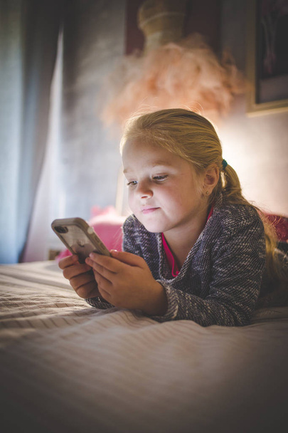 Beautiful blond toddler girl laying on her bed watching videos on a smart phone - Foto, afbeelding