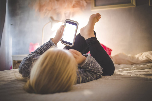Beautiful blond toddler girl laying on her bed watching videos on a smart phone - Photo, image