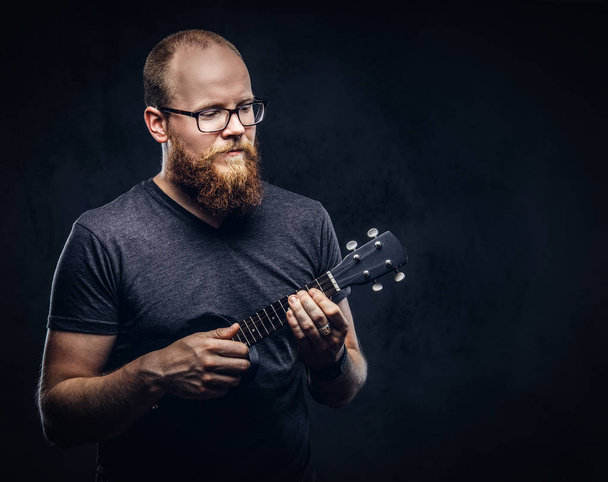 Roodharige gebaarde mannelijke musicus bril gekleed in een grijs t-shirt op een ukulele spelen. Geïsoleerd op donkere gestructureerde achtergrond.  - Foto, afbeelding