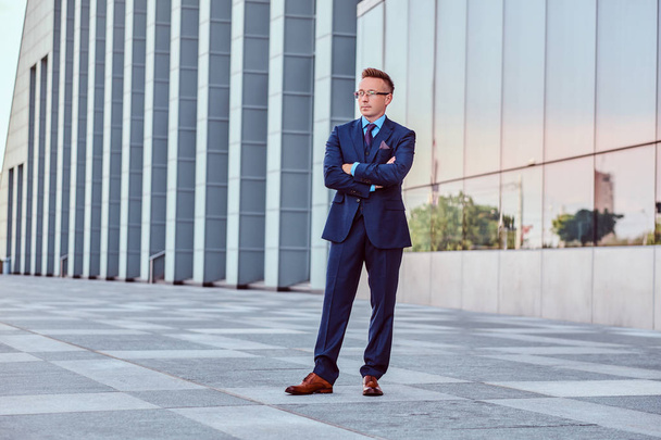 Confident businessman dressed in elegant suit standing with crossed arms against cityscape background. - Photo, Image