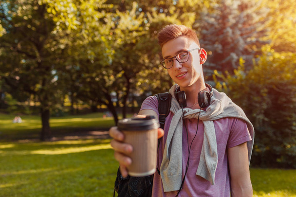 Handsome college guy chilling in spring campus park. Happy man student wears earphones and glasses and drinking coffee - Фото, изображение