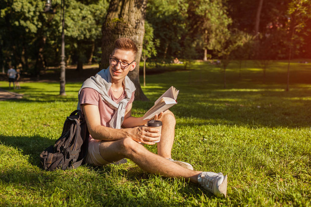 Handsome college man reading a book and drinking coffee in campus park. Happy guy student learning outdoors sitting on grass in spring. Education concept - Photo, Image