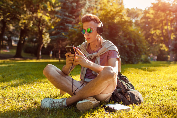 Beau collégien accroché au smartphone et écoutant de la musique dans le parc du campus. heureux guy étudiant relaxant assis sur herbe en été
 - Photo, image
