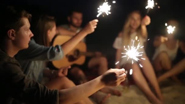 Друзья на пляже с бенгальскими огнями, празднующими закат. Молодые люди сидят у костра с бенгальскими огнями и поют. Бородатый хипстер играет на гитаре
. - Кадры, видео