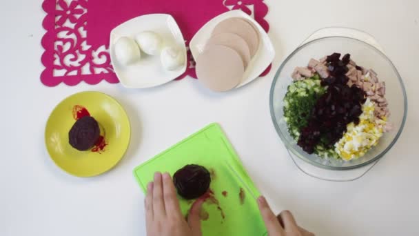 The cook cuts the boiled beets on a cutting board. Nearby in the dishes are other ingredients for cooking sugar beet, chicken egg, sausage and cucumber. View from above. - Video