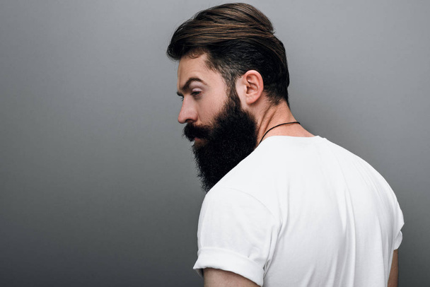 Rear view of brutal attractive young bearded male model looking aside, posing on a gray studio background. Portrait of handsome young European fashion barber with trendy beard. - 写真・画像