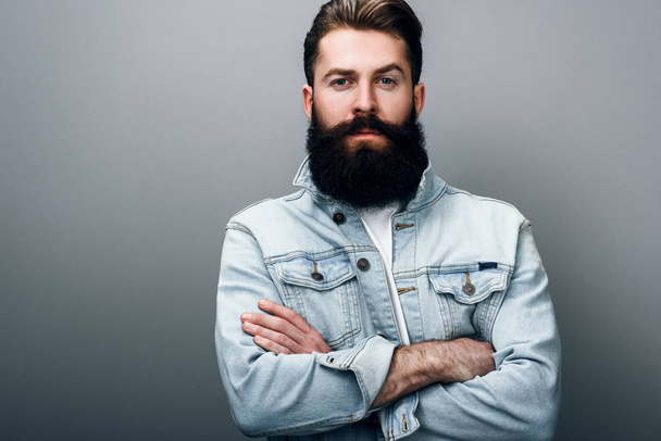 Brutal handsome fashionable young European male with thick beard wearing trendy denim jacket, cross hands, staring at camera with serious and cheerful look. Barber man posing against gray studio wall. - Zdjęcie, obraz