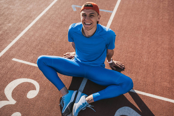 Happy smiling Caucasian athlete male doing stretching exercises sitting on the running track. Runner sitting on running track with feet joined together preparing for a run or relaxing after workout. - Photo, Image