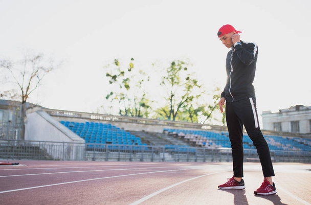 handsome young athletic male wearing earphones looking down to the track in the stadium. Attractive runner listening to music during morning jog and run on outdoor. Sport, people and lifestyle. - Foto, Imagen