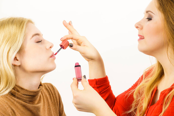 Make up artist friend applying another woman lipstick or lip gloss on lips. Two friends interested in visage. - Foto, imagen