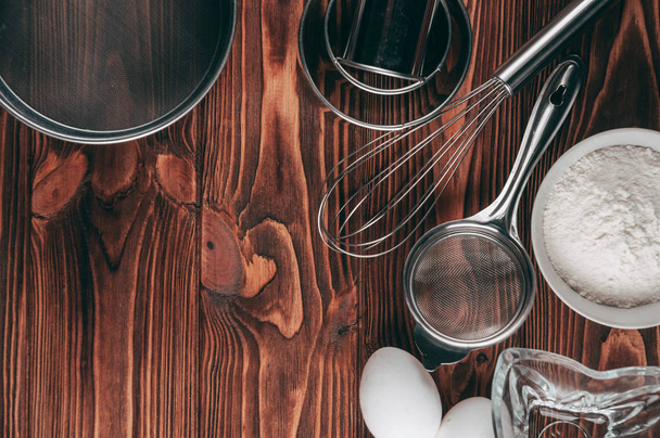 Ingredients in the dishes on a wooden brown table, where as well lie the whisk is full for flour, three chicken eggs and a slate board. View from above. Place under the text - Foto, Imagem