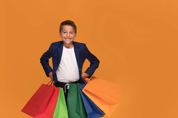 Little boy in a business suit with colorful shopping bags posing on orange studio background, smiling. - Foto, Imagem