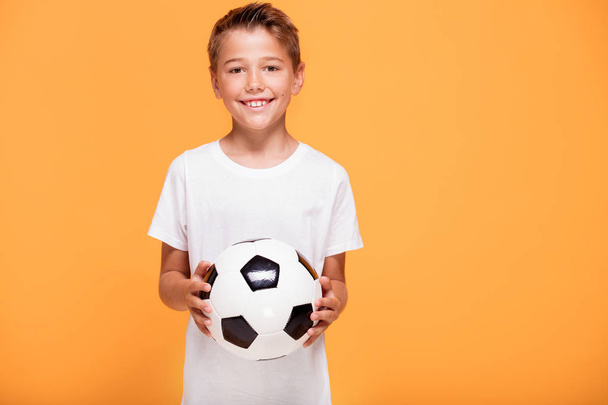 Happy little player hold soccer ball, smiling to the camera. A lot of copy space , yellow studio background. - Fotografie, Obrázek