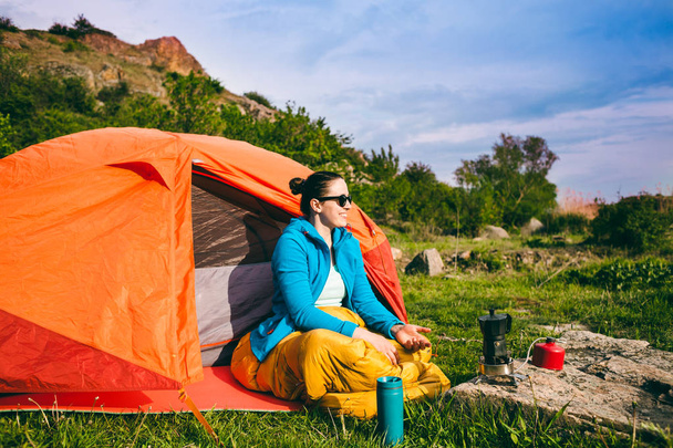 Zelten im Wald. Ein Mädchen bereitet Kaffee auf einer Geysir-Kaffeemaschine zu. eine Frau sitzt in einem Schlafsack neben einem Zelt und bereitet vor dem Hintergrund der Berge Kaffee zu. - Foto, Bild