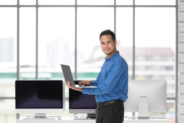 Young Asiabn businessman standing and holdin new laptop computer with happiness manner in modern office, group of desktop computers blur in behide. - Photo, Image