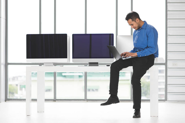 Jeune homme d'affaires Asiabn assis et tenant un nouvel ordinateur portable avec bonheur manière dans le bureau moderne, groupe d'ordinateurs de bureau flou dans behide
. - Photo, image