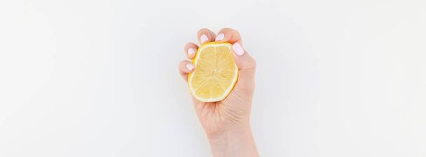 パステル カラーのマニキュア光沢ホワイト バック グラウンド コピー スペース ミニマリズム スタイルに分離されたレモンの半分を保持している女性の手。フェミニンなソーシャル メディアの長い広いバナー。健康的な食事の概念 - 写真・画像