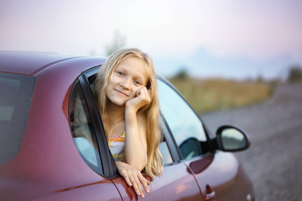 gelukkig glimlachend meisje kijken uit de auto raam en zonsondergang aan de achterzijde - Foto, afbeelding