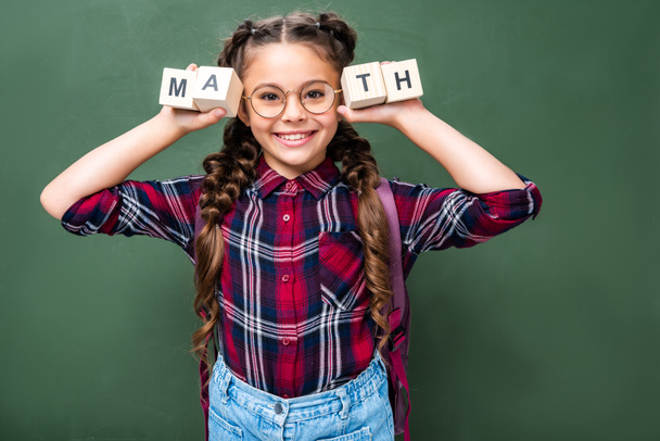 smiling schoolchild holding wooden cubes with word math near blackboard - Photo, Image