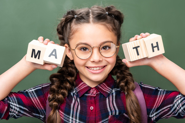 portrait of schoolchild holding wooden cubes with word math near blackboard - Foto, immagini
