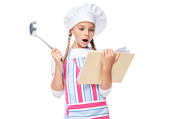 shocked schoolchild in costume of chef reading cookbook isolated on white - Фото, изображение