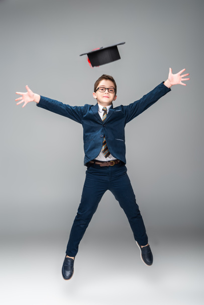 schoolboy in businessman costume jumping with graduation cap isolated on grey - Photo, Image