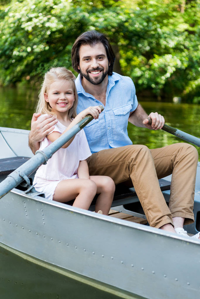 glücklicher Vater und Tochter beim Bootsfahren und Blick auf die Kamera auf dem See im Park - Foto, Bild