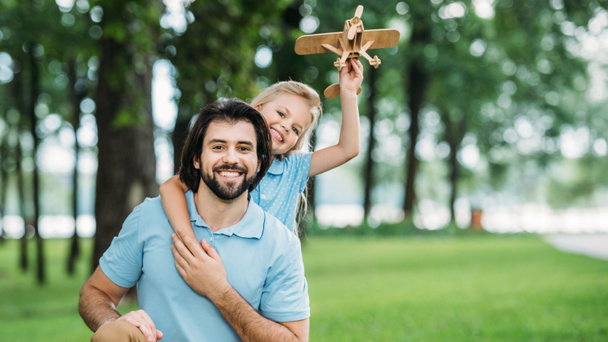 adorable little daughter embracing happy father from behind and holding toy airplane at park - Photo, Image