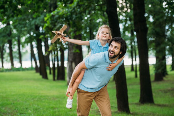 lachende dochter meeliften op gelukkige vader en spelen met speelgoed vliegtuig in park - Foto, afbeelding