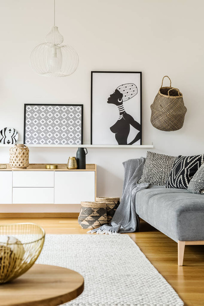 Real photo of a couch with cushions and blanket standing next to a cupboard in a boho living room interior with white rug, posters on a shelf and table in the front - Valokuva, kuva