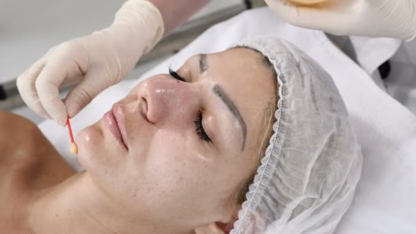 Beauty concept. Young female client gets beauty facial procedure. face renovation. Beautician uses cotton bud to cover face skin with beauty mask - Footage, Video