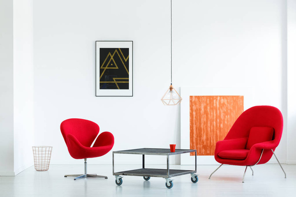 Real photo of two red armchairs standing next to a metal table with wheels in minimal white living room interior with two posters and a lamp - Foto, Bild