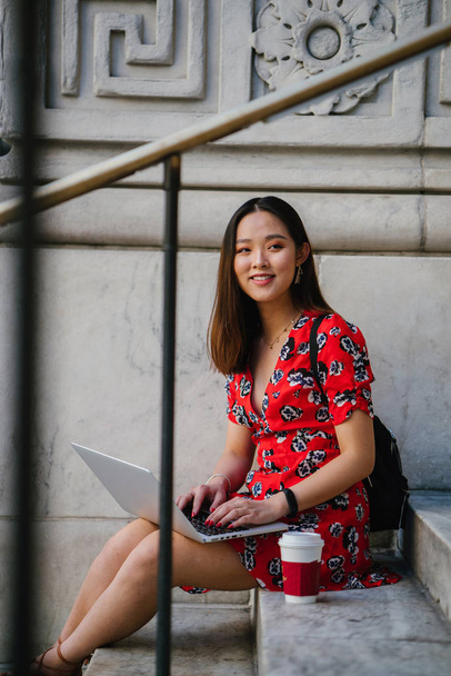Asian woman in red dress working on laptop sitting on steps - Fotoğraf, Görsel