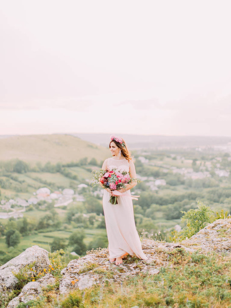 The full-length view of the bride with the wedding bouquet looking aside at the background of the mountains. - Foto, afbeelding