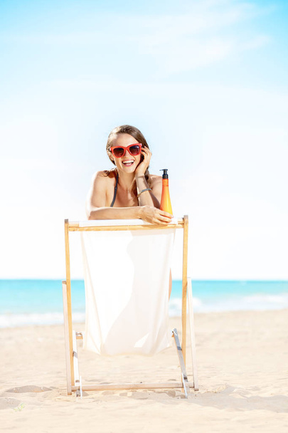 happy modern woman in swimwear on the seashore with suntan lotion sitting in a beach chair - Foto, Imagem