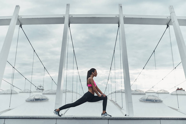 sportive woman stretching outdoors on roof of modern building against cloudy sky  - Foto, Imagen