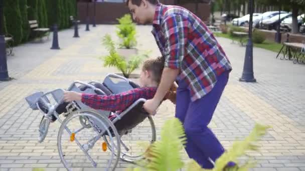 Man rolls a happy teenager in a wheelchair in a summer Park. - Πλάνα, βίντεο