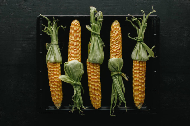 vista superior de mazorcas de maíz crudas frescas en bandeja para hornear negra sobre mesa de madera
 - Foto, imagen