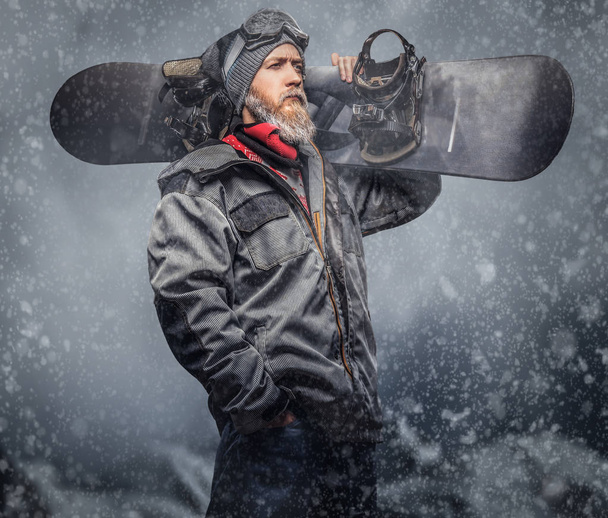 Brutální zrzka snowboardista s plnovousem v zimní čepice a ochranné brýle sobě snowboarding kabát pózuje s snowboard na pozadí hor. - Fotografie, Obrázek