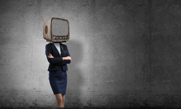 Business woman in suit with old TV instead of head keeping arms crossed while standing inside empty room with gray dark wall on background. - Foto, immagini